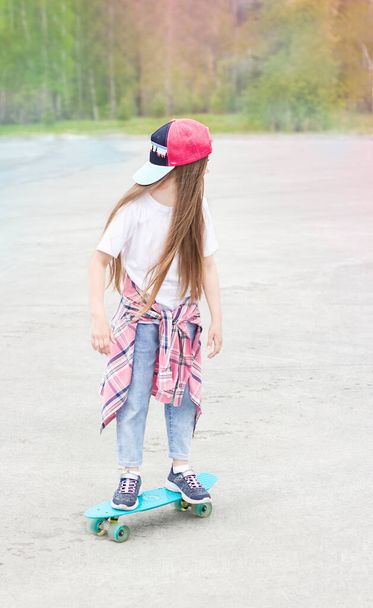 little smiling girl stands outdoors with a skateboard in her hands on the background of the forest - Фото, изображение