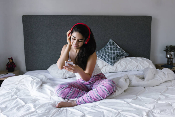 Young latin woman listening music with headphones on bed at home in Mexico Latin America - Photo, image
