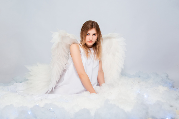 An attractive girl in a white tunic with large white wings behind her back poses while sitting in white luminous clouds. - Fotoğraf, Görsel
