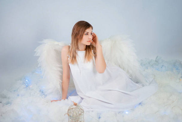 An attractive girl in a white tunic with large white wings behind her back poses while sitting in white luminous clouds. - Valokuva, kuva