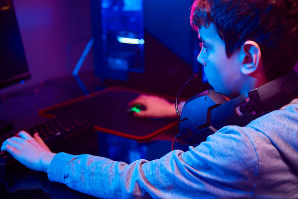 Мальчик играет в компьютерную видеоигру в темной комнате, использует неоновую цветную механическую клавиатуру RGB, рабочее место для киберспортивных игр, детскую игровую зависимость - Фото, изображение
