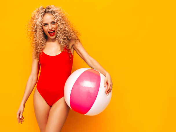 Young beautiful smiling woman posing near yellow wall in studio.Sexy model in red swimwear bathing suit.Positive female with curls hairstyle. Holding penny inflatable ball.Happy and cheerful - 写真・画像