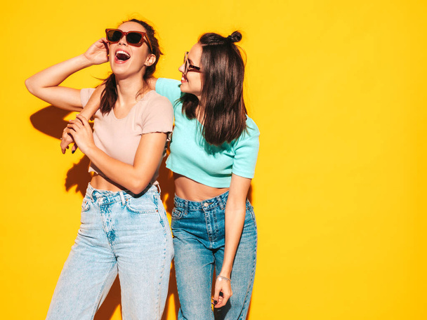 Two young beautiful smiling blond hipster female in trendy summer clothes. Sexy carefree women posing near yellow wall in studio. Positive models having fun. Cheerful and happy. In sunglasses - Foto, imagen