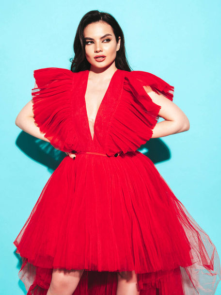 High fashion portrait of young beautiful brunette woman wearing nice trendy red evening fluffy dress. Sexy fashion model posing in studio. Fashionable female isolated on blue - Φωτογραφία, εικόνα