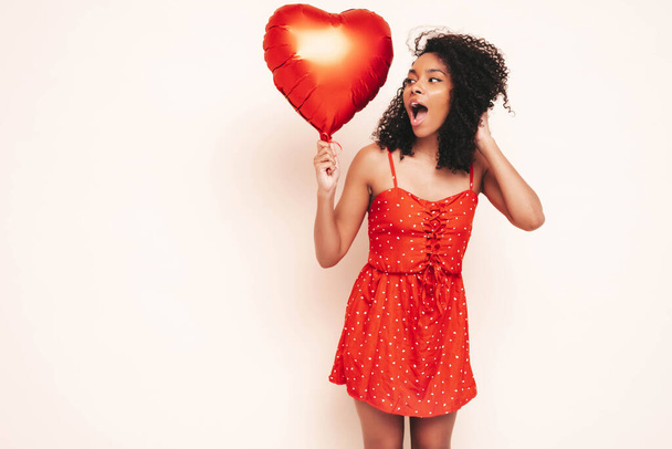 Beautiful black woman with afro curls hairstyle. Smiling model dressed in red summer dress. Sexy carefree female posing near wall in studio. Tanned and cheerful.Holding heart air balloon.Isolated - Foto, afbeelding
