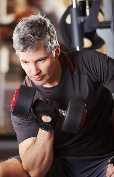 Definiendo sus bíceps. Un hombre maduro haciendo rizos de bíceps con una mancuerna en el gimnasio. - Foto, Imagen