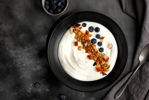 Greek yogurt with Granola with coconut, apple pieces in bowl. Breakfast with muesli, yoghurt and blueberries, dark background - Foto, immagini