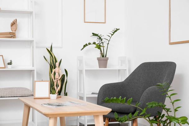 Cómodo sillón, mesa, plantas de interior y estantería cerca de la pared blanca - Foto, Imagen