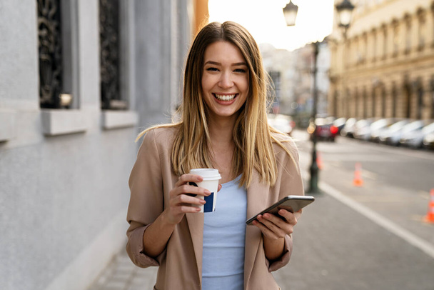 Attractive young successful woman drinking a coffee and using a smartphone while enjoying break - Photo, Image
