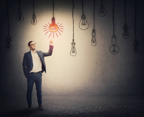 Cheerful businessman points up index finger, showing an enlightened light bulb among multiple others switched off. Business metaphor, idea choice concept - Photo, Image