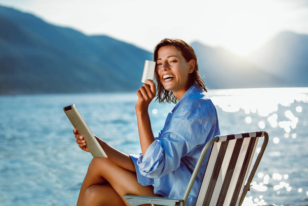 Mujer feliz con tableta digital y tarjeta de crédito comprar en línea sentado cerca del mar. - Foto, Imagen
