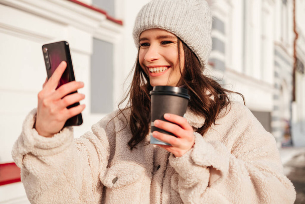 Caucasian smiling young woman in warm coat drinking coffee and taking selfie with mobile phone outdoors - Fotoğraf, Görsel