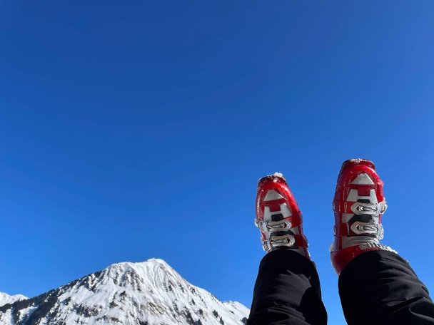 Ноги жінки в червоних лижних чоботах відпочивають після лижного туру в сонячний день, сніжні Альпи на задньому плані. Фотографія високої якості - Фото, зображення