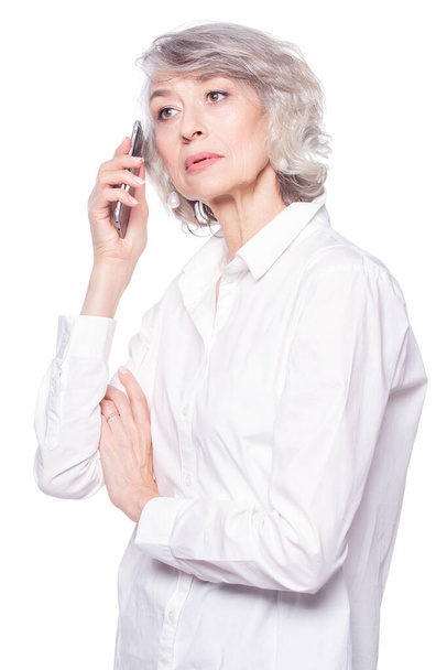 An attractive elderly woman listens very attentively to the interlocutor during a telephone conversation using a smartphone isolated on white background - Фото, зображення