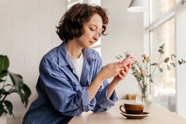 Молода жінка, що сидить у кафе, користується мобільним телефоном і п'є каву. Цариця з кучерявим коротким волоссям у приміщенні. - Фото, зображення