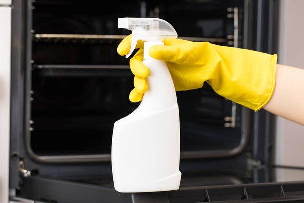  The cleaner holds a mockup of an oven detergent spray bottle in front of the oven in a yellow household glove.  - Zdjęcie, obraz