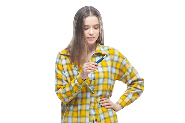 Portrait of a young woman in a yellow checkered shirt looking at electronic cigarette in her hands isolated on white background - Photo, image