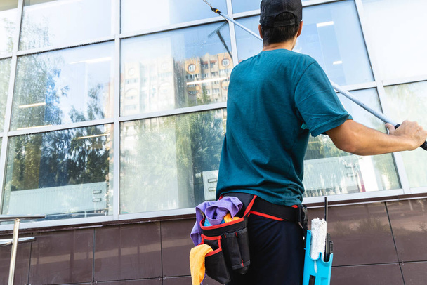A young male worker of a professional cleaning service in overalls washes the glass of the windows of the facade of the building. Clean showcases on request - 写真・画像