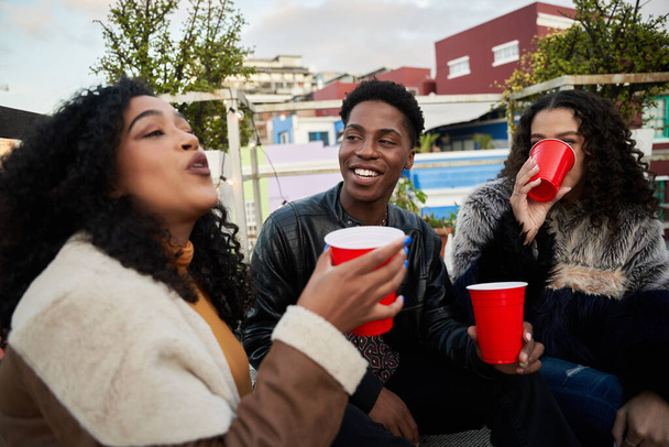 Group of multi-cultural young adults sitting on a rooftop terrace socializing, drinking and laughing - Fotó, kép