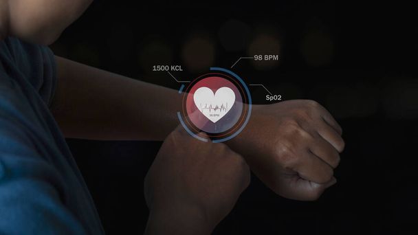 Akıllı saat teknolojisi kullanan adam ekrandaki sağlık uygulaması simgesiyle kalp atışlarını kontrol ediyor. Holografik simge kullanıcı arayüzü. Gelecekçi akıllı saat teknolojisi. Sağlık hizmetleri kavramı. - Fotoğraf, Görsel