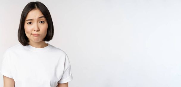 Imagen de joven morena asiática en camiseta blanca, sonrisa y mirada con duda, vacilante, de pie sobre fondo blanco - Foto, imagen