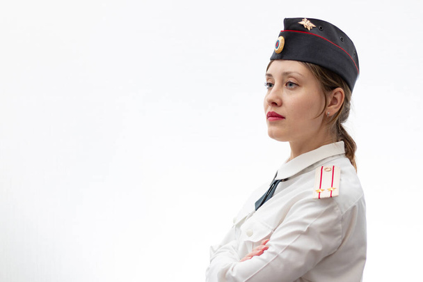 Una hermosa joven oficial de policía rusa vestida de uniforme con una gorra y tirantes de un teniente coronel y una camisa blanca sobre un fondo blanco. Enfoque selectivo. Retrato - Foto, imagen