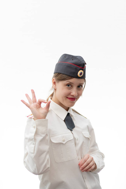 Hermosa joven oficial de policía rusa en uniforme de vestir muestra señales de mano perfectamente y sonríe sobre un fondo blanco. Enfoque selectivo. Retrato - Foto, imagen