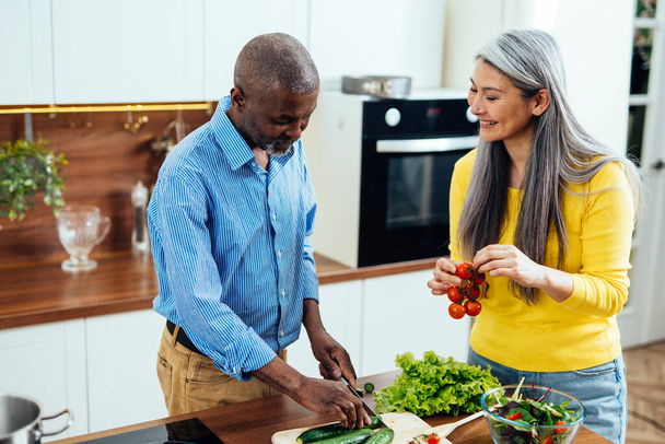 cinematic image of a multiethnic senior couple preparing food in the kitchen. Indoors Lifestyle moments at home. Concept about seniority and relationships - Fotó, kép