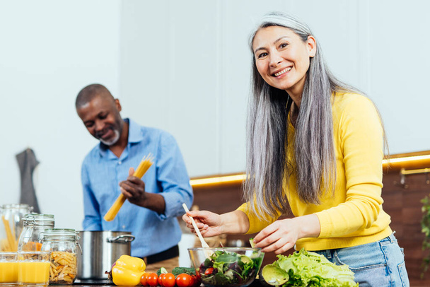 cinematic image of a multiethnic senior couple preparing food in the kitchen. Indoors Lifestyle moments at home. Concept about seniority and relationships - Fotografie, Obrázek
