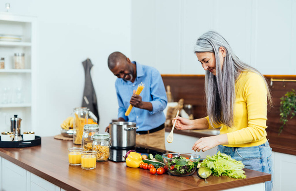 cinematic image of a multiethnic senior couple preparing food in the kitchen. Indoors Lifestyle moments at home. Concept about seniority and relationships - Foto, immagini
