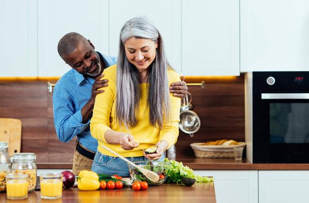 cinematic image of a multiethnic senior couple preparing food in the kitchen. Indoors Lifestyle moments at home. Concept about seniority and relationships - Foto, afbeelding