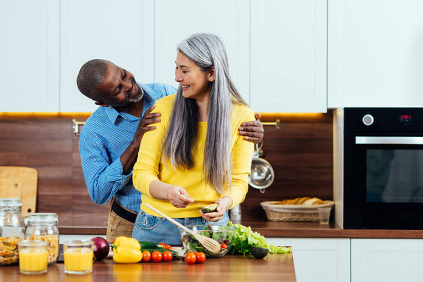 cinematic image of a multiethnic senior couple preparing food in the kitchen. Indoors Lifestyle moments at home. Concept about seniority and relationships - Valokuva, kuva