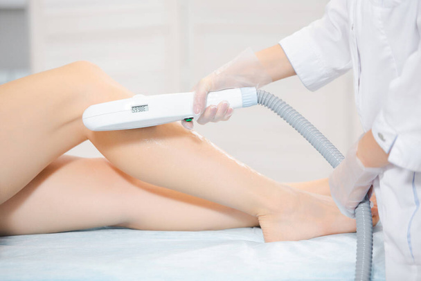 Close-up das pernas do paciente deitado no salão de beleza e as mãos esteticista realizando o procedimento de depilação a laser com instrumentos modernos - Foto, Imagem