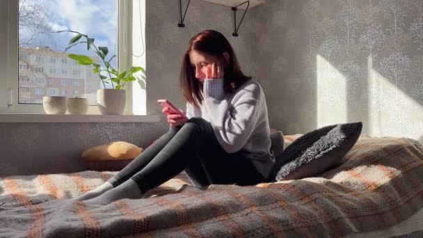 Yatakta sosyal medyada gezinmek için akıllı telefon kullanan rahat genç bir kadın.. - Video, Çekim