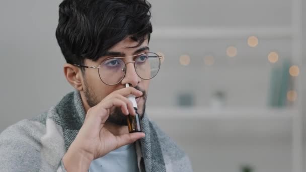 Indian bearded sad man sick at home using antibacterial spray for runny nose sighs tired looking at camera frustrated sickness covid seasonal flu cold guy in glasses wrapped in blanket suffering virus - Footage, Video