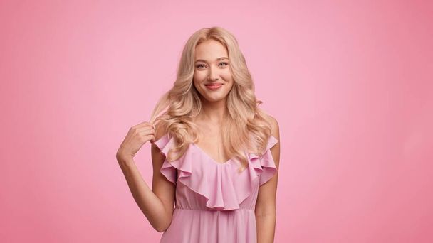 Πανέμορφη κυρία παίζει με κατσαρά ξανθά μαλλιά πάνω από ροζ φόντο - Φωτογραφία, εικόνα