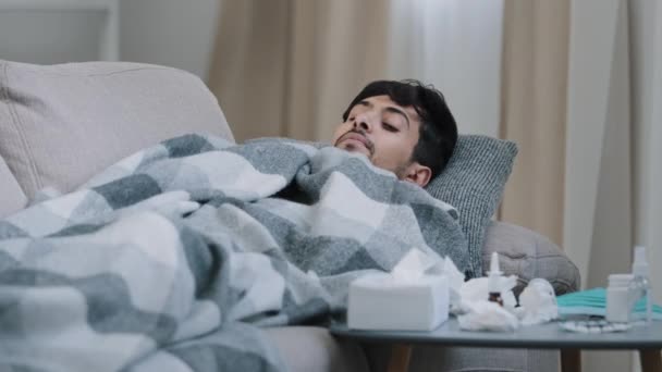 Indian bearded sad exhausted man lying on home couch ill covered blanket suffering from fever disease symptoms coronavirus concept looking at medicines on table tired of treating covid19 seasonal flu - Footage, Video
