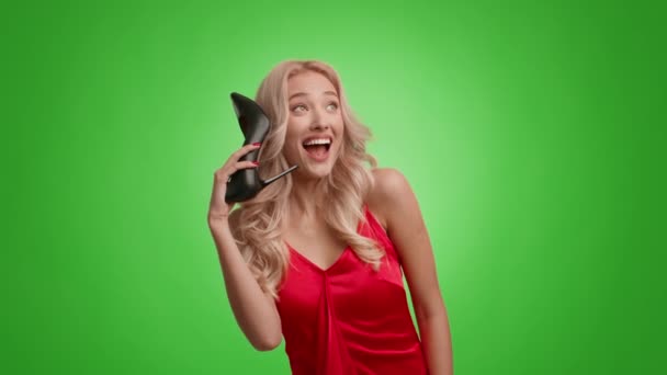 Блондинка, яка робить вигляд, що розмовляє по телефону Холдинг взуття, зелений фон - Кадри, відео
