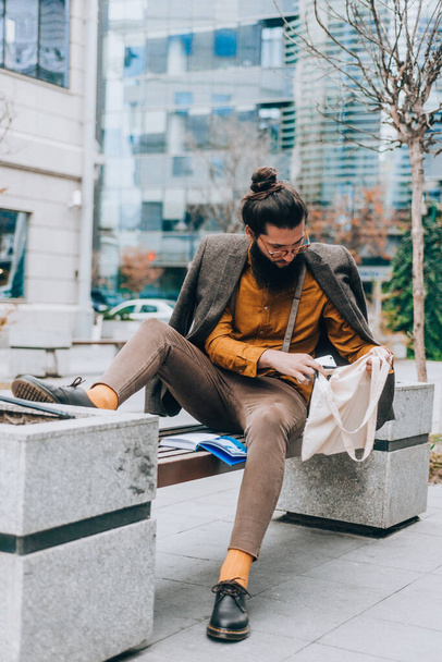 Σύγχρονη hipster νεαρός άνδρας κρατώντας μια τσάντα, ενώ κάθεται στον πάγκο στην πλατεία της πόλης - Φωτογραφία, εικόνα