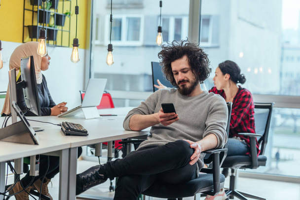 Bearded employee guy with afro haircut talking on his phone while being surrounded by his multiethnic coworkers. Businessman working in a casual startup modern environment. - Foto, immagini