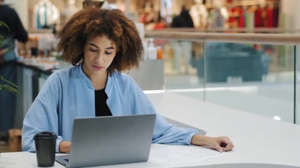Африканская американская занятая деловая женщина с ноутбуком в офисе кафе на рабочем месте компании проверяет данные финансирования бюджета записи в документах, делая заметки, работая с компьютером дистанционного электронного обучения - Кадры, видео