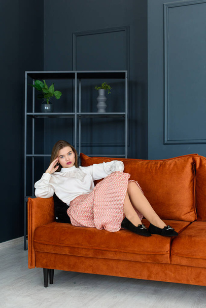 Divatos fiatal nő portréja bézs szoknyában, fehér blúzban és elegáns bézs színű fekete Seude cipőben, csattal, narancssárga kanapén pózolva. - Fotó, kép