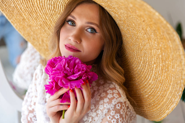Close-up portrait of a beautiful woman in a straw hat, looking at the camera. Woman with makeup and red lips, red peony in her hands. Blonde with curled hair - Foto, afbeelding