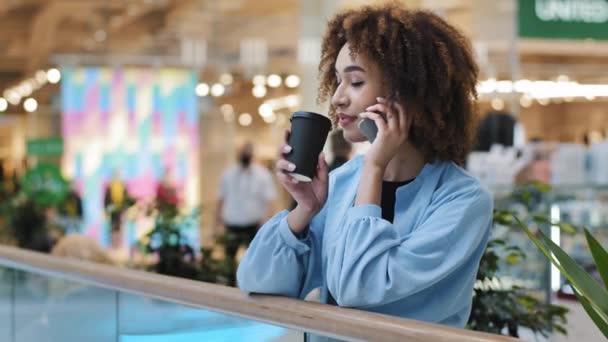 Female african american business woman talking phone distant friendly conversation call calling online booking making order with smartphone girl drinking tea coffee in mall company mobile tech talk - Footage, Video