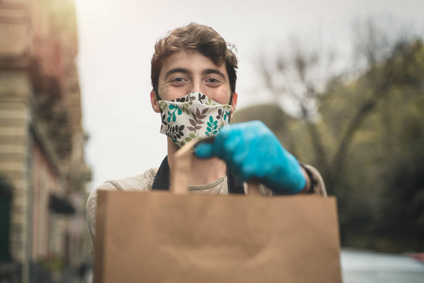 The delivery boy shows the package with the takeaway food holding it with gloves and smiling through the coronavirus protective mask - Photo, Image