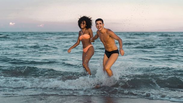 海の上で手をつないでビーチでリラックスして楽しんでいる異人種間の若いカップルの完全な長さの肖像画。夏の楽しいコンセプト. - 写真・画像