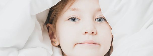 Little girl under white blanket. The warmth of home comfort - Valokuva, kuva