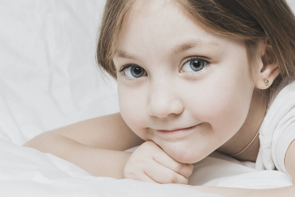 Beautiful little girl is lying on bed with white linens. The warmth of home comfort - Φωτογραφία, εικόνα