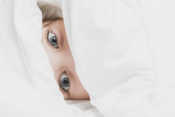 Beautiful eyes of little girl under white blanket. The warmth of home comfort - Φωτογραφία, εικόνα