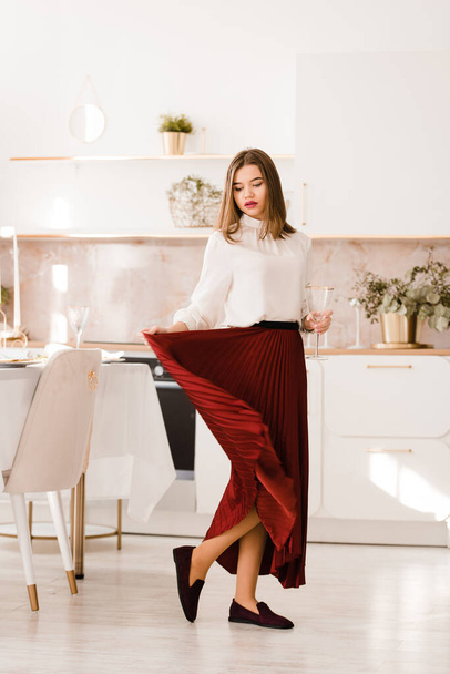 Portrait of fashionable woman in a red skirt, white blouse and stylish suede shoes with a buckle posing on the kitchen. Girl with a vine glass in a hands - Φωτογραφία, εικόνα